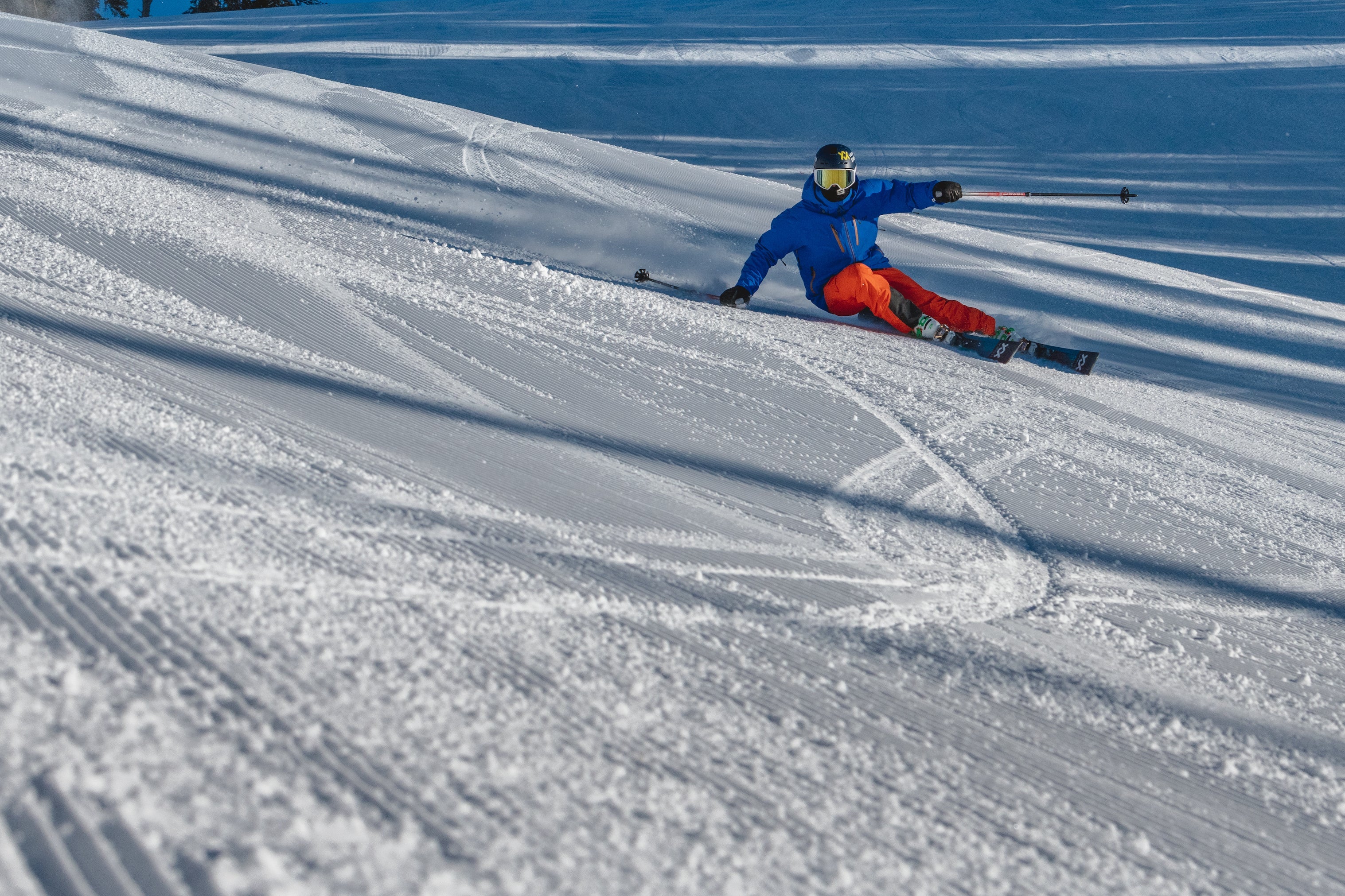 Skiier in blue jacket and red pants skiing down a snowy slope with ski tracks.