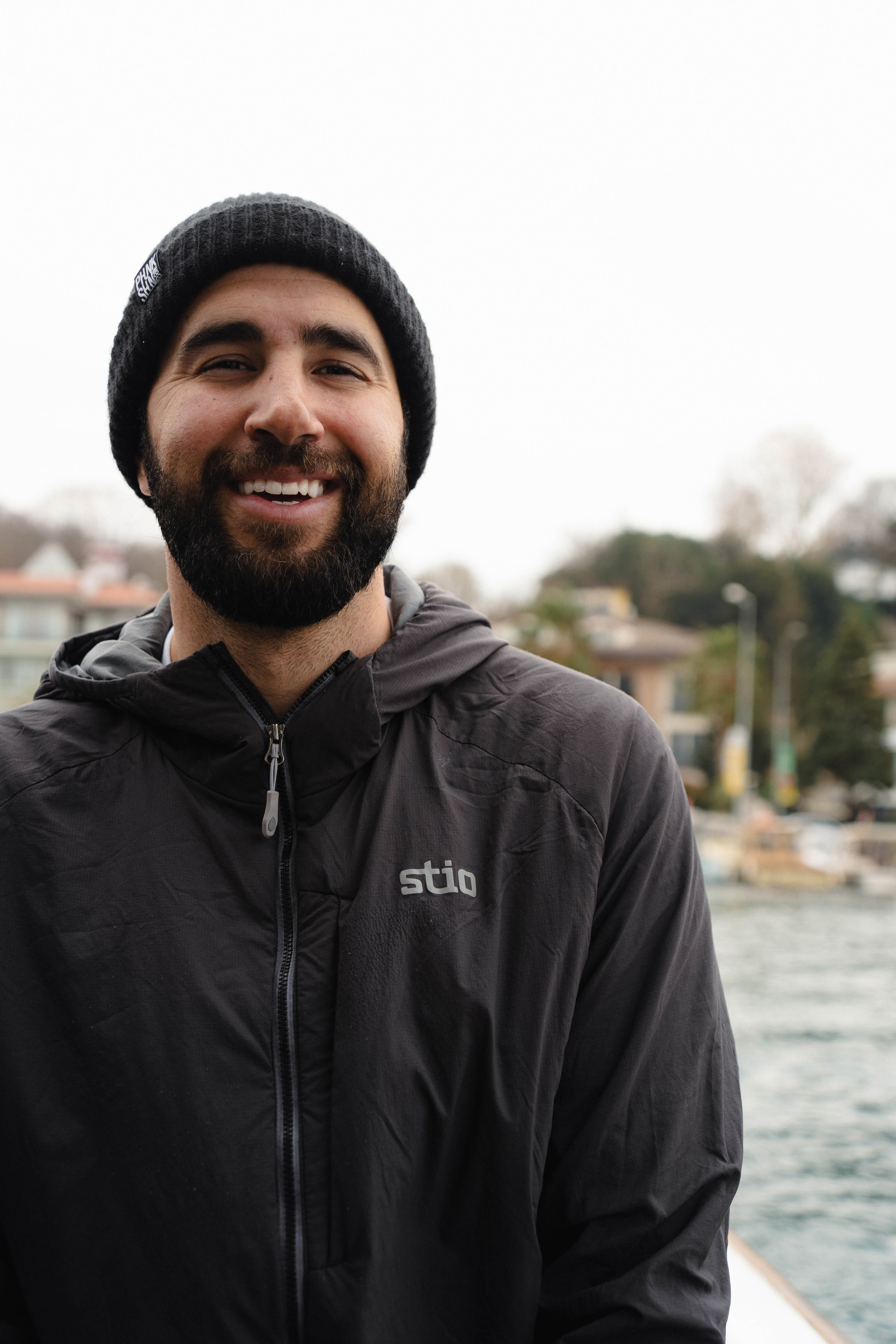 Man wearing a black jacket with 'Stio' logo, standing by a body of water.